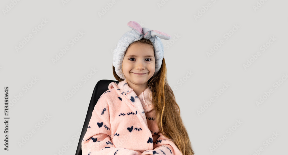 Beautiful little girl with blond hair, isolated on a white background, dressed in a pink robe and with a blue headband in her hair, is ready for procedures at the salon. Fashion concept. Beautiful little girl with blond hair, isolated on a white background, dressed in a pink robe and with a blue headband in her hair, is ready for procedures at the salon. Fashion concept.