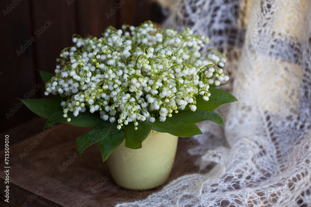 Bouquet of spring lily of the valley flowers in a vase on a wooden chair, white lace shawl, blur, still life.