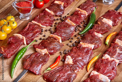 Metal skewers with raw uncooked beef meat for frying on the wooden board with peppers, spices and cherry tomatoes near it, close-up perspective view shallow depth of field. Meat and spices in focus
