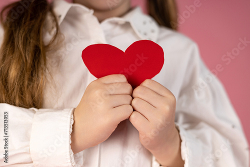 A little girl's hands holding a red paper heart to her chest on a pastel pink background. Valentine's day concept