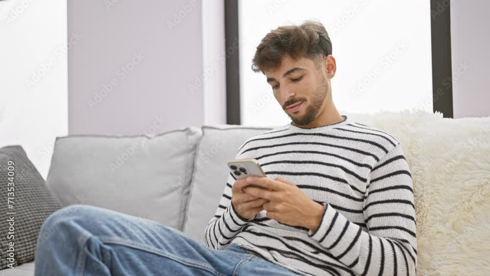 Cheerful young arab man joyfully texting on his smartphone while sitting comfortably on his sofa, exuding happiness in the warm, cozy living room of his home Cheerful young arab man joyfully texting on his smartphone while sitting comfortably on his sofa, exuding happiness in the warm, cozy living room of his home
