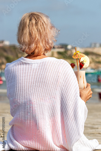 Toma cercana de mujer mediana edad con pelo pintado de rosa sentada de espalda en la playa con un cocktail en mano relajada, descansando del trabajo photo