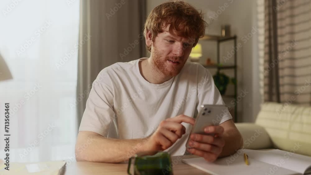 Handsome young red haired man browse products in internet store on smartphone while finds promotional sale offer and immediately reached for credit card starting to make purchase order online at home
