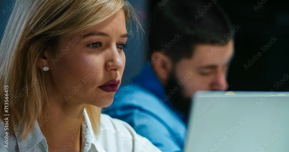 Close up of beautiful blonde young woman working at laptop computer in office late at night. Pretty female company worker typing and looking at screen. Male colleague on blurred background. Close up of beautiful blonde young woman working at laptop computer in office late at night. Pretty female company worker typing and looking at screen. Male colleague on blurred background.