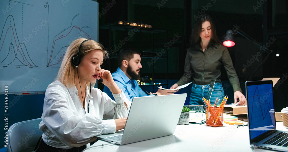Caucasian young attractive woman in headphones talking in call-center office and working on laptop. Female call center worker typing on computer while speaking with client. Colleagues on background. Caucasian young attractive woman in headphones talking in call-center office and working on laptop. Female call center worker typing on computer while speaking with client. Colleagues on background.