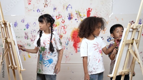 Diverse student painted or draw canvas at stained wall in art lesson. Asian girl wearing white shirt with stained color while standing at stained wall with hand print. Creative activity. Erudition.