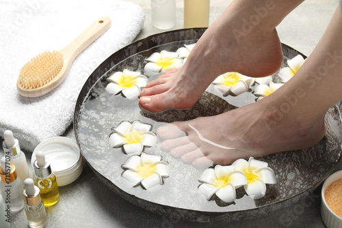Woman soaking her feet in bowl with water and flowers on light grey floor, closeup. Spa treatment