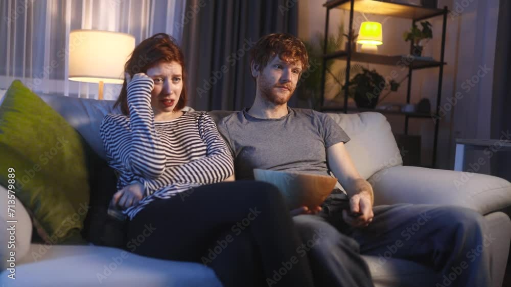 Tired young red haired family couple with remote controller switches channels on TV trying to choose something interesting looking at screen at late night Boring TV shows films movie Tired young red haired family couple with remote controller switches channels on TV trying to choose something interesting looking at screen at late night Boring TV shows films movie