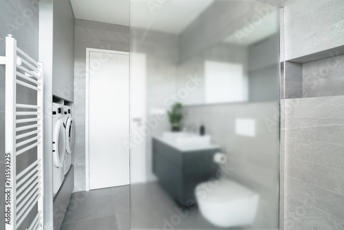 A small bathroom with a toilet, sink, washing machine and dryer. The walls and the floor are made of gray tiles. There is a big milky glass wall, which separates shower from the rest of the bathroom. photo