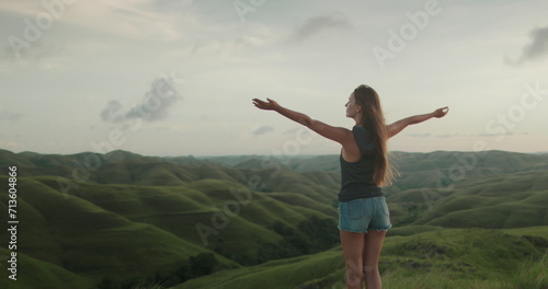 Woman stand on green hill meadow, raised hands, wind blow her hair - freedom, enjoy nature. Tourist girl watch sunset beautiful landscape. Outdoor lifestyle travel summer holiday vacation.