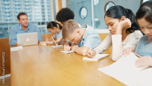 Young smart teenager writing and talking about topic at classroom. Diverse highschool student taking a note in other notebook while sitting with multicultural friends. Creative education. Edification.