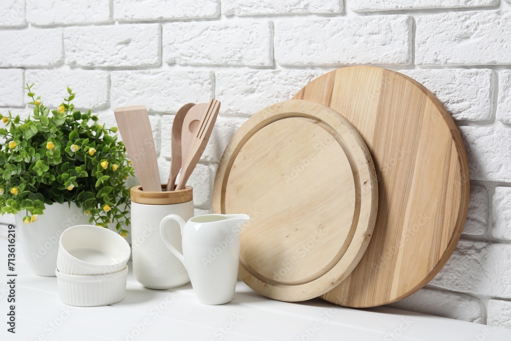 Cutting boards, holder with kitchen utensils, dishware and houseplant on white table near brick wall Cutting boards, holder with kitchen utensils, dishware and houseplant on white table near brick wall