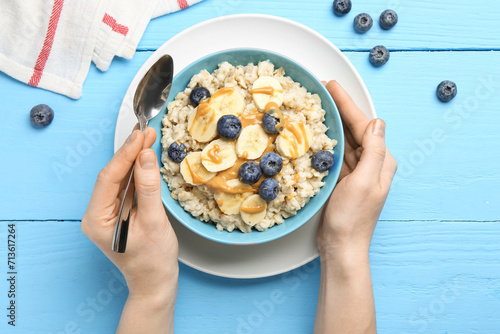 Woman eating tasty oatmeal with banana, blueberries and peanut butter at light blue wooden table, top view