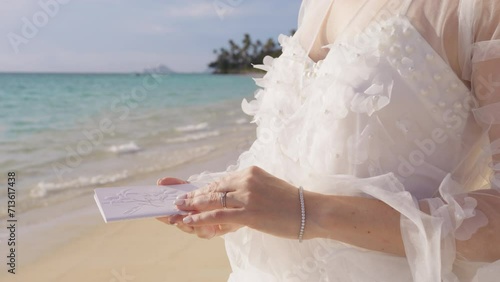 Unrecognizable bride in beautiful white floral boho wedding dress saying her vows to groom at beach wedding ceremony. Closeup shot of female hands holding embossed vow books. Elopement background