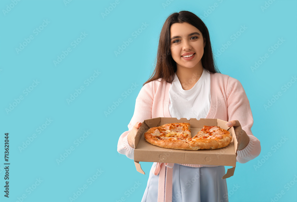 Young woman holding cardboard box with tasty pizza on blue background