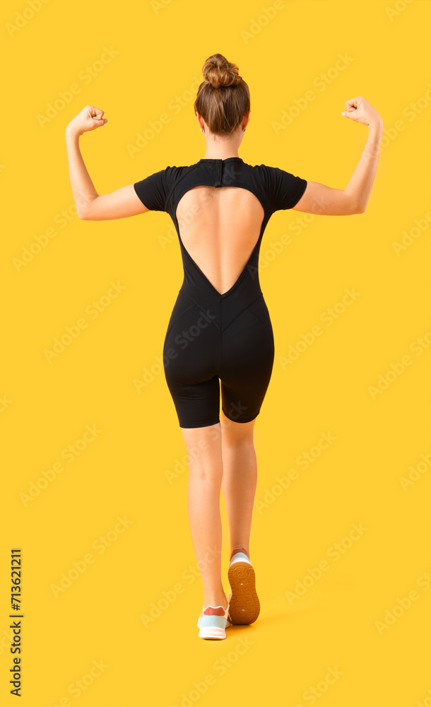 Young woman in cycling jumpsuit showing muscles on yellow background, back view Young woman in cycling jumpsuit showing muscles on yellow background, back view