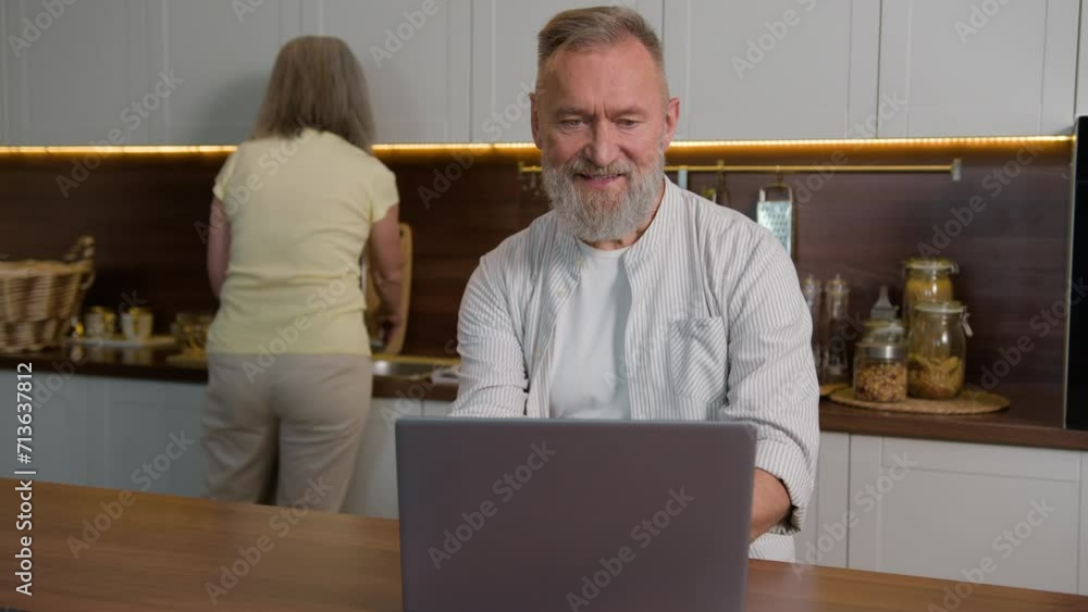 Smiling Caucasian man retired adult mid aged businessman husband typing on laptop computer working online shopping internet store pay bills at home while mature woman wife housewife cook in kitchen Smiling Caucasian man retired adult mid aged businessman husband typing on laptop computer working online shopping internet store pay bills at home while mature woman wife housewife cook in kitchen