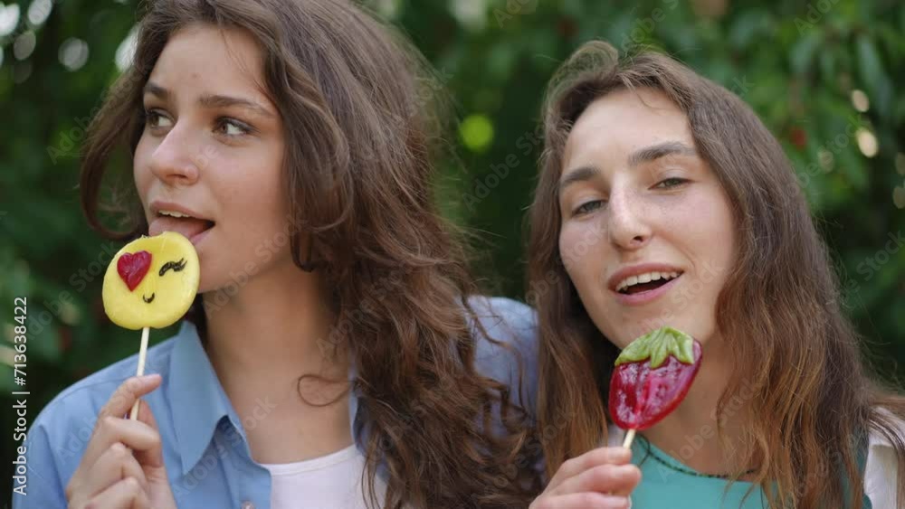 Close-up. Girls in shirts eat lollipops while standing outdoors. The girls laugh and exchange candy Close-up. Girls in shirts eat lollipops while standing outdoors. The girls laugh and exchange candy
