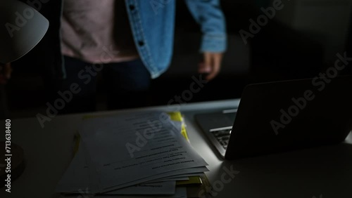 A focused man works overtime on documents and a laptop in a dimly lit office, highlighting dedication and late hours. photo