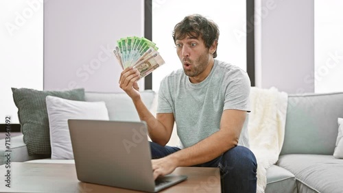 Shocked young man at home, face displaying disbelief, mouth open in amazement, handling russian rubles on laptop, expressing fear and surprise! photo