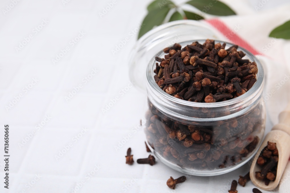 Aromatic cloves in glass jar and scoop on white tiled table, closeup. Space for text Aromatic cloves in glass jar and scoop on white tiled table, closeup. Space for text