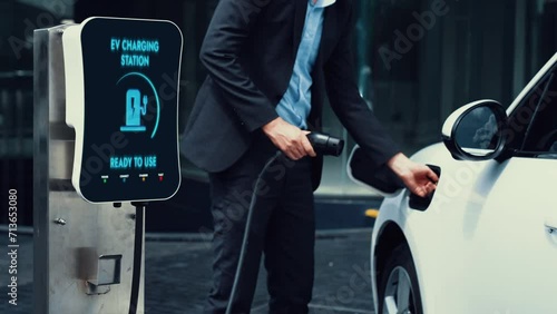 Businessman insert EV charger to recharge her electric car from EV charging station at city center or public car park. Eco friendly rechargeable car using alternative energy in city lifestyle.Peruse photo