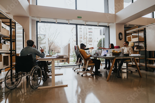 A multiracial group of employees collaborates around a table in a well-lit office space, showcasing wheelchair accessibility and teamwork. © qunica.com