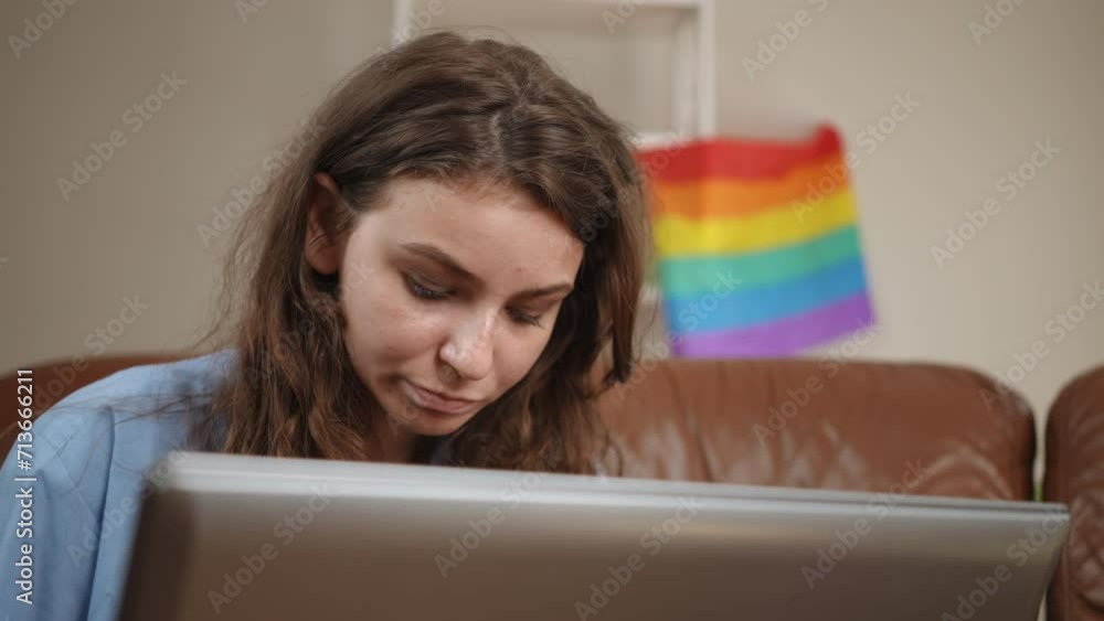 Close-up. A girl with long hair is concentrating on typing on a laptop keyboard while sitting on a leather sofa in a spacious living room with a rainbow flag on the wall Close-up. A girl with long hair is concentrating on typing on a laptop keyboard while sitting on a leather sofa in a spacious living room with a rainbow flag on the wall