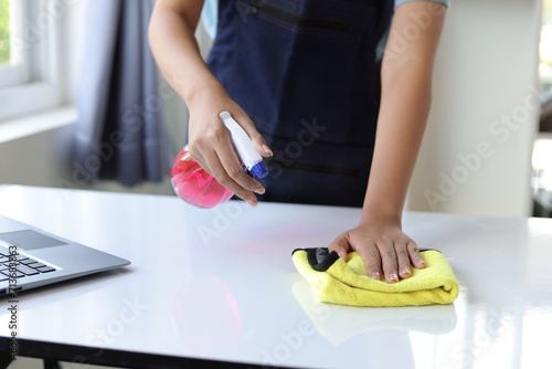 A young woman wearing an apron is cleaning a table in her home office using disinfectant and a wipe.