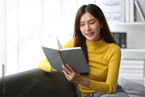 Charming Asian woman is happily reading a book on the sofa in the living room at home.