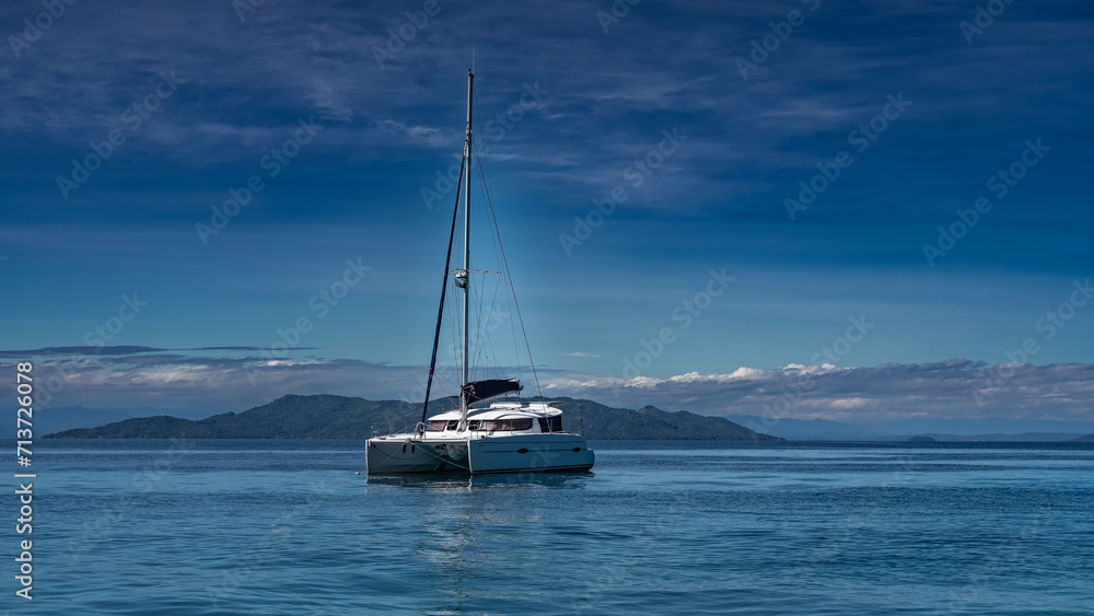 The white yacht is anchored in the ocean. A tall mast against a background of blue sky and clouds. Mountains in the distance. Ripples and highlights on the water. Madagascar. Nosy Tanikeli The white yacht is anchored in the ocean. A tall mast against a background of blue sky and clouds. Mountains in the distance. Ripples and highlights on the water. Madagascar. Nosy Tanikeli