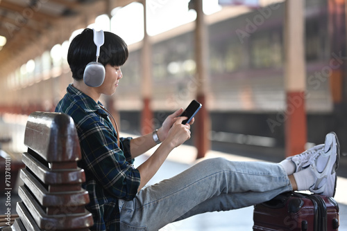 Young man in headphones using mobile phone while sitting on bench at railway station photo