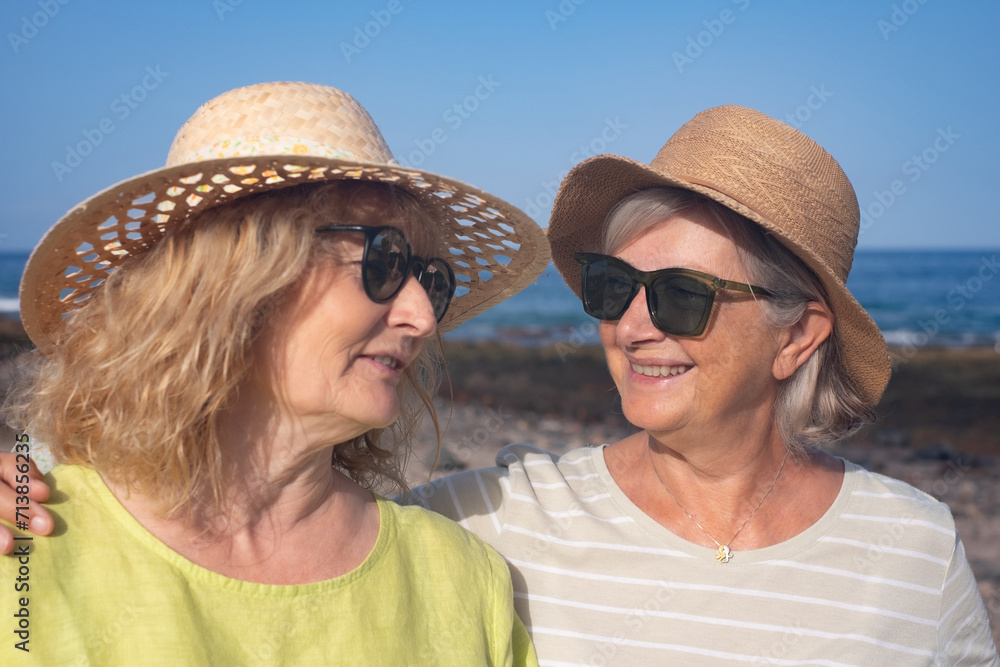 Happy couple of senior and middle aged women embraced standing outdoors at sea wearing sunglasses and hats. Vacation, freedom, friendship concept. Horizon over water