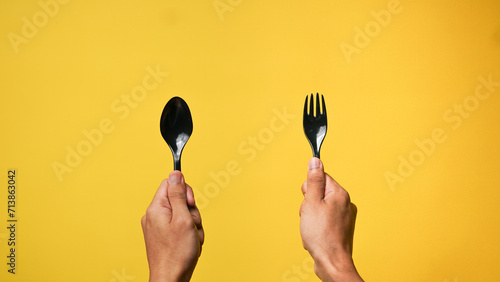 Man's hand holding a black spoon and fork on a yellow background