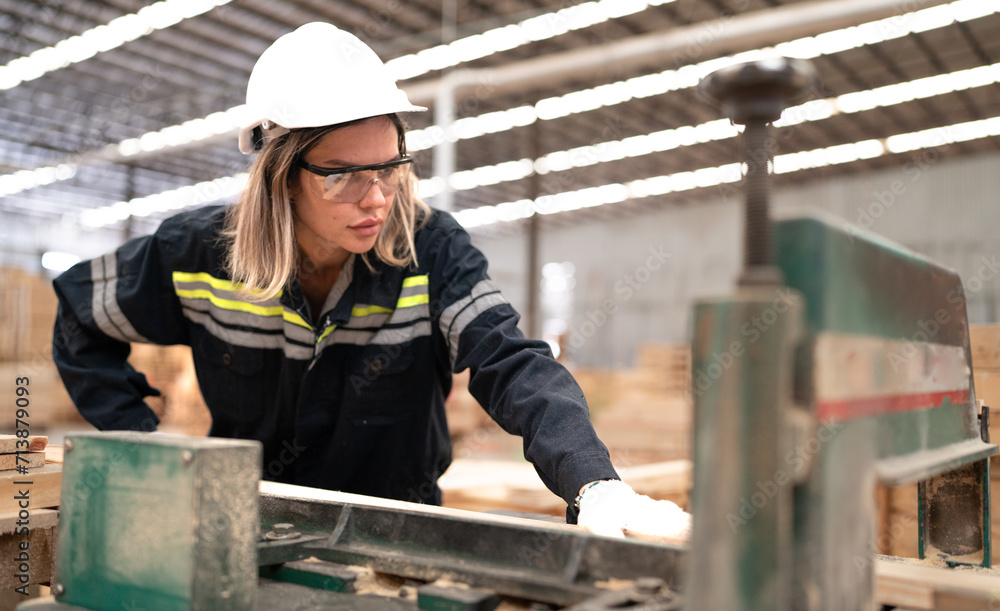 Confident female worker working in hardwood warehouse of wooden furniture factory checking stock. Busy skilled inspector in uniform hardhat examining plank pallet material for production facility.