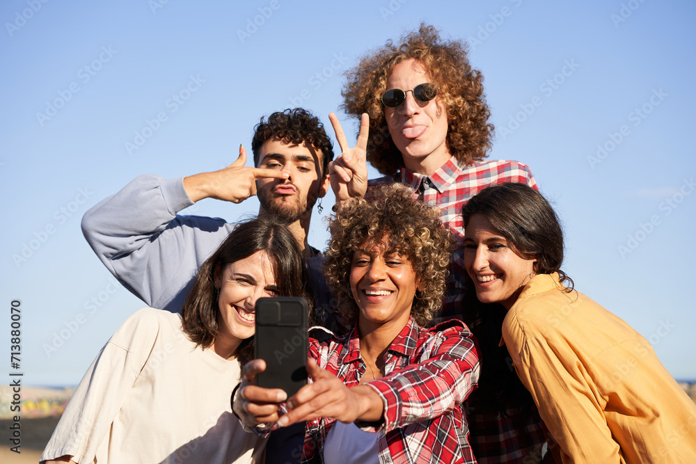 Young adult group of diverse cheerful friends making funny selfie phone. Millennial multiracial people using mobile for smiling photo sunny day outdoor. Concept of community and positive relationship. Young adult group of diverse cheerful friends making funny selfie phone. Millennial multiracial people using mobile for smiling photo sunny day outdoor. Concept of community and positive relationship.