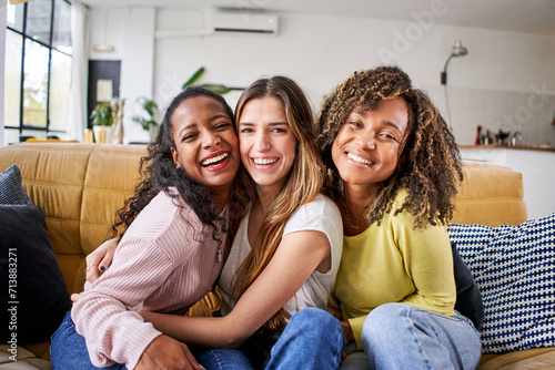 Portrait of three attractive female room mates looking smiling at camera. Group of cute cheerful girls hugging sitting on couch at home. Happy friends posing for photo indoors. Generation z and rent. photo