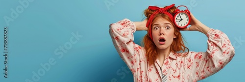 Anxious, panicking young redhead girl missed interview, overslept, holding red alarm clock and touching head frustrated with concerned face, wearing nightwear and sleep mask, blue background.  photo