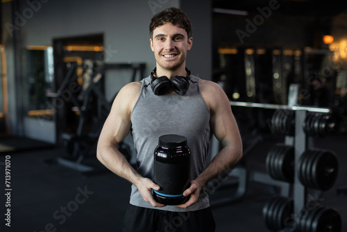 Smiling man athlete posing at gym with container of protein whey, holding pack with fitness supplements, muscular guy in sport club, place for your design