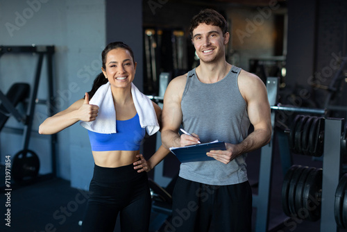 Happy young woman showing thumb up, male personal trainer holding clipboard, standing in gym and smiling at camera