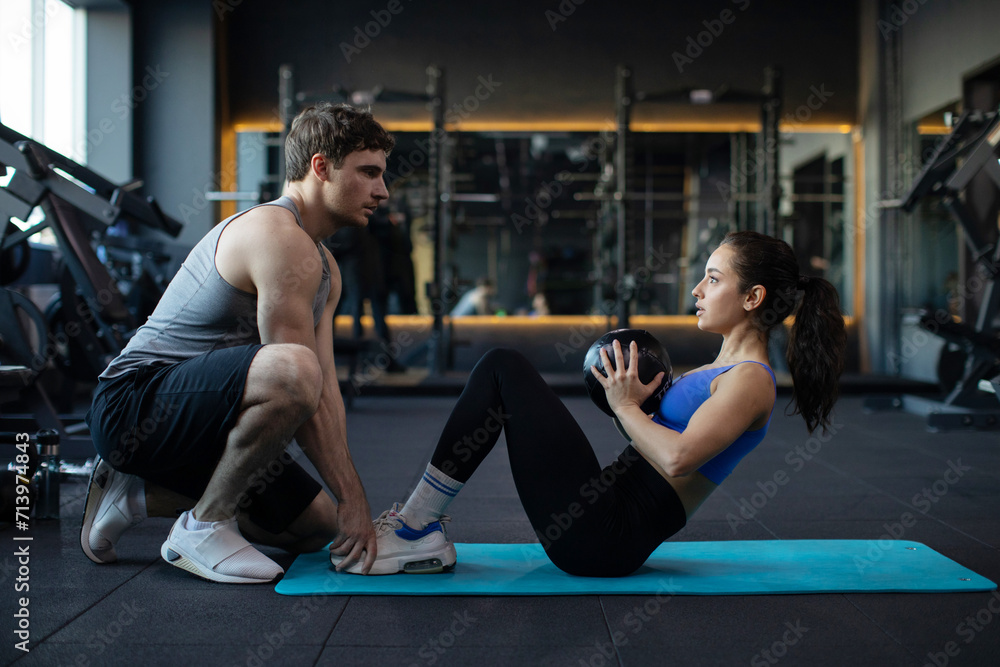 Sporty young couple exercising together in gym, man assisting fit lady while doing sits ups with ball in fitness centre, side view Sporty young couple exercising together in gym, man assisting fit lady while doing sits ups with ball in fitness centre, side view