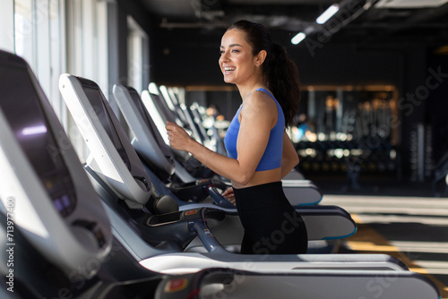 Happy young european woman running on treadmill in modern gym, smiling and radiating positivity, enjoying morning fitness routine