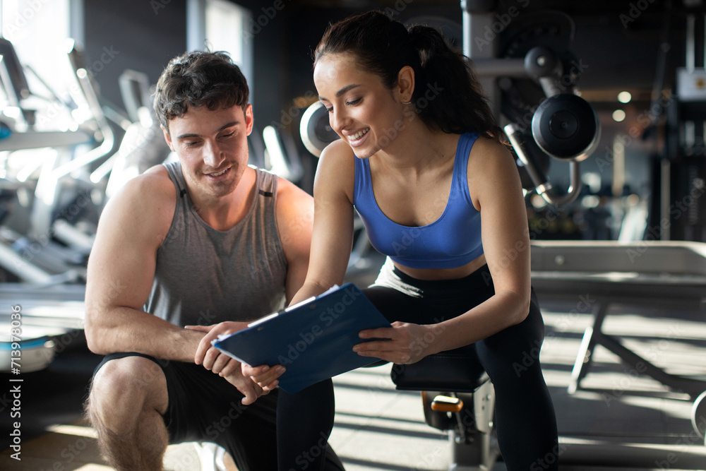 Fototapeta Female personal trainer and her male client discussing nutrition or training plan on clipboard, sitting in modern gym interior. Healthy lifestyle, fitness concept
