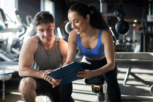 Female personal trainer and her male client discussing nutrition or training plan on clipboard, sitting in modern gym interior. Healthy lifestyle, fitness concept