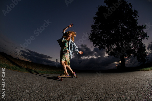 Young smiling girl in a dress and a denim jacket is riding a longboard on an asphalt road