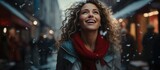 A vibrant woman beams with joy on a chilly street, her curly hair and red scarf adding a pop of color to her winter jacket