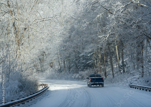 A lone pickup truck drives through a wintry mountain landscape on a snow-covered road. The road curves ahead with guardrails on both sides. 