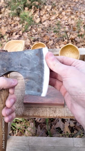 Close-up of a carpenter checks the sharpness of an axe and starts to sharpen it. He sits on a camping chair in a forest.