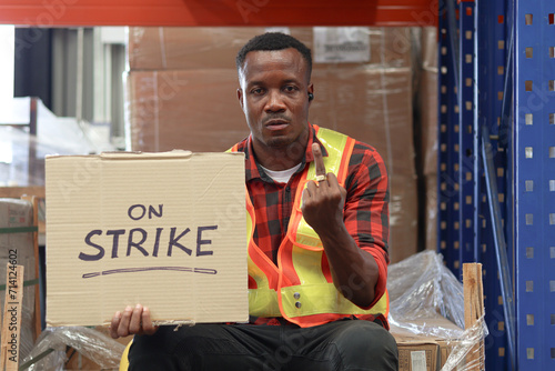 Angry unhappy African worker man wearing safety vest and giving the middle finger with strike banner placard sign at cargo logistic warehouse. Striking worker protesting at workplace. photo