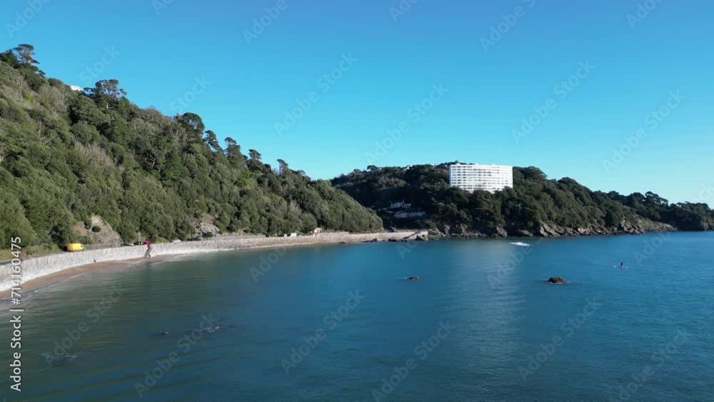 Meadfoot beach, Torbay, South Devon, England: DRONE VIEWS: Wide angle view of the beach, offshore rocks, a large apartment block overlooking the sea and a paddleboarder at sea. Meadfoot beach, Torbay, South Devon, England: DRONE VIEWS: Wide angle view of the beach, offshore rocks, a large apartment block overlooking the sea and a paddleboarder at sea.